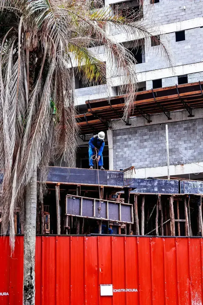 Trabalhador de construção civil de capacete branco e uniforme azul manuseando corda em obra, com palmeira e tapume vermelho
